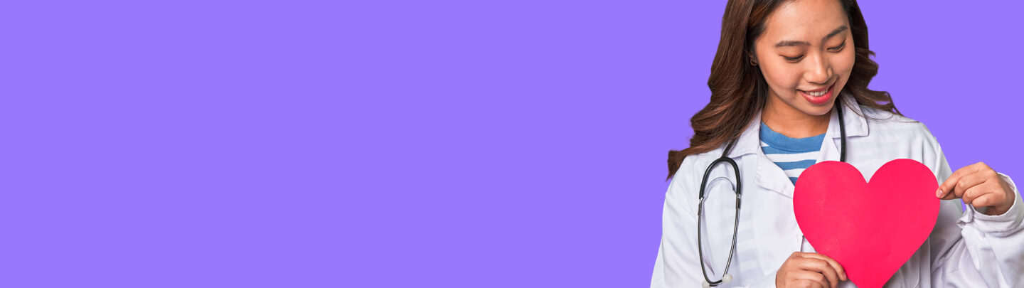 Smiling doctor in a white coat holding a red paper heart against a purple background.